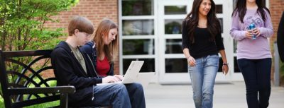 Two students sitting on outdoor bench with laptops while two smiling students walk by.