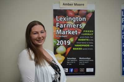 Smiling Student stands in front of Lexington Farmers Market poster