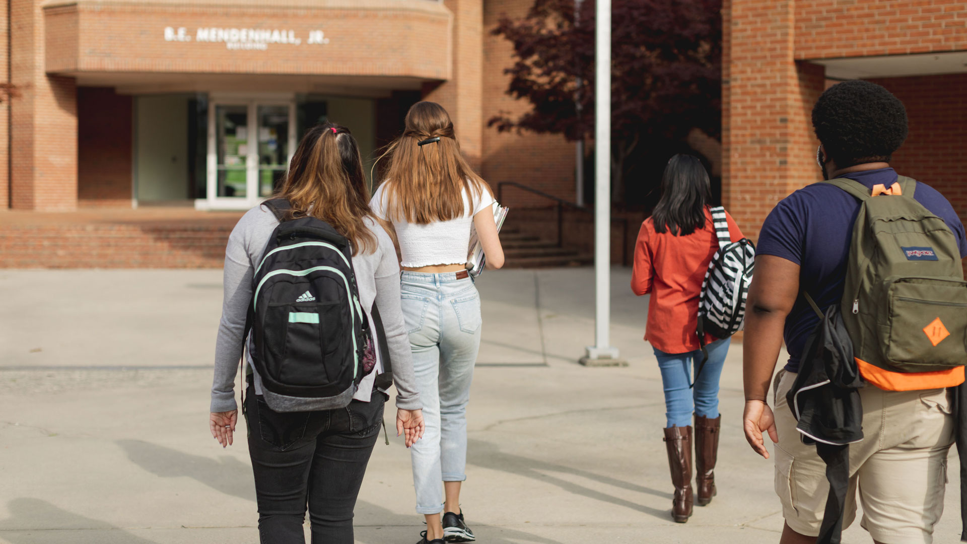 Students with book bags walking away from camera toward bricked building