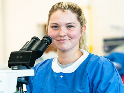 Student in scrubs smiles at camera while sitting in front of a microscope