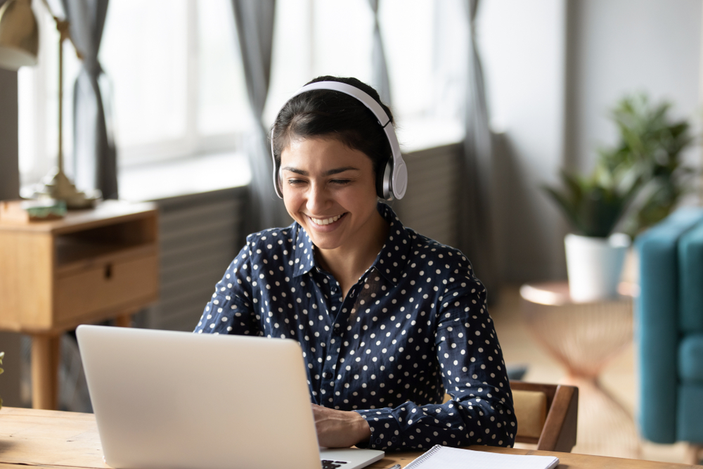 Smiling female in polkadot shirt wearing headphones and sitting in front of laptop.
