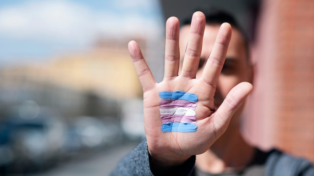 closeup of the palm of the hand of a person with a transgender flag painted in it, in front of their face