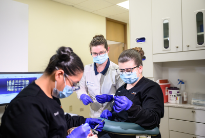 Three dental professionals in a clinic, two performing a procedure, one teaching.