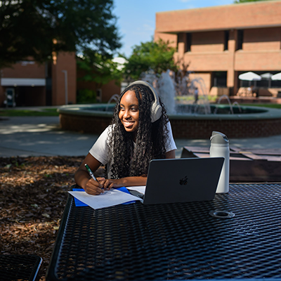 A woman with headphones writing at an outdoor table with a laptop and a fountain in the background.