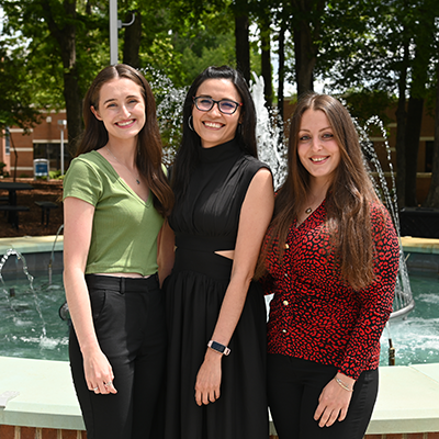 Three women standing in front of a fountain, smiling on a sunny day.