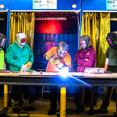 People in a workshop wearing welding gear, with one actively welding.
