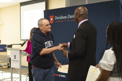 Two men shaking hands at a community college event, one in a navy hoodie, the other in a black suit.