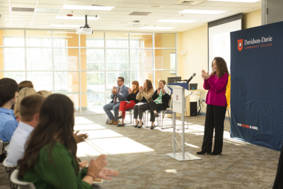A woman claps at a podium in a bright room with a seated panel and audience, banners, and large windows.